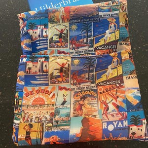 Könnte beinhalten: Ein Buch mit dem Titel "Elin Hilderbrand" ragt aus einer farbenfrohen Buchhülle heraus. Die Hülle ist mit Vintage-Reisepostern in Blau-, Orange- und Weißtönen gemustert, die ein Küstenmotiv hervorrufen. Das Design enthält Bilder der französischen Riviera.