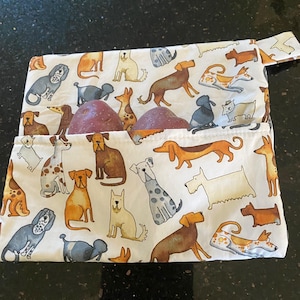 Puede incluir: Una bolsa de tela blanca con un patrón repetitivo de varias razas de perros en tonos marrón, gris y naranja. La bolsa tiene un bolsillo frontal que contiene dos patatas rojas enteras. El diseño incluye múltiples ilustraciones caninas.