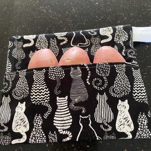 Può includere: Un sacchetto di stoffa nero e bianco a tema gatto che contiene tre patate rosse. La borsa presenta un motivo ripetuto di gatti stilizzati. Le patate sono di colore rosa-rossastro.