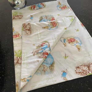 May include: A white cotton fabric with a repeating pattern of Peter Rabbit characters in blue, red, and brown. The fabric is folded in half, showing the pattern on both sides.