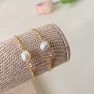Pu&ograve; includere: Due braccialetti a catena dorata, ognuno con un'unica perla bianca. I braccialetti sono esposti su un supporto in tessuto beige.
