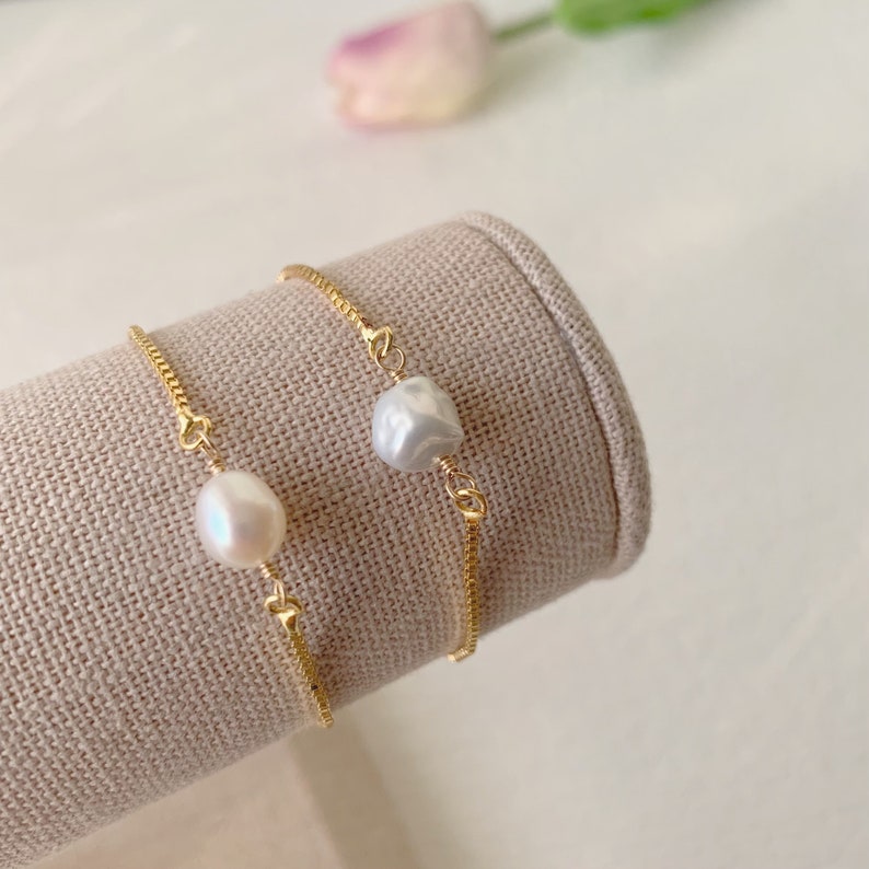 Pu&ograve; includere: Due braccialetti a catena dorata, ognuno con un'unica perla bianca. I braccialetti sono esposti su un supporto in tessuto beige.