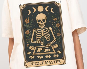 May include: Cream-colored t-shirt featuring a graphic of a skeleton assembling a puzzle, framed by floral elements and celestial bodies. The text "PUZZLE MASTER" is at the bottom of the design. The shirt has short sleeves and a relaxed fit.