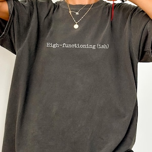 Peut inclure: T-shirt gris foncé surdimensionné avec le texte "High-functioning (ish)" en blanc. Le t-shirt a des manches courtes et une coupe décontractée. Le tissu semble doux et légèrement délavé.