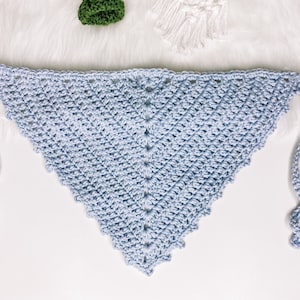 Puede incluir: Una bufanda triangular de ganchillo de color azul claro con un borde festoneado y dos lazos largos.