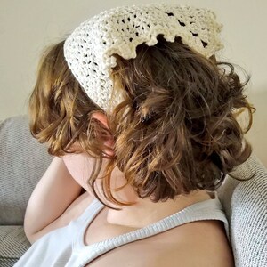 Puede incluir: Una diadema de crochet blanca con un borde festoneado. La diadema est&aacute; puesta en una persona con el pelo rizado marr&oacute;n.