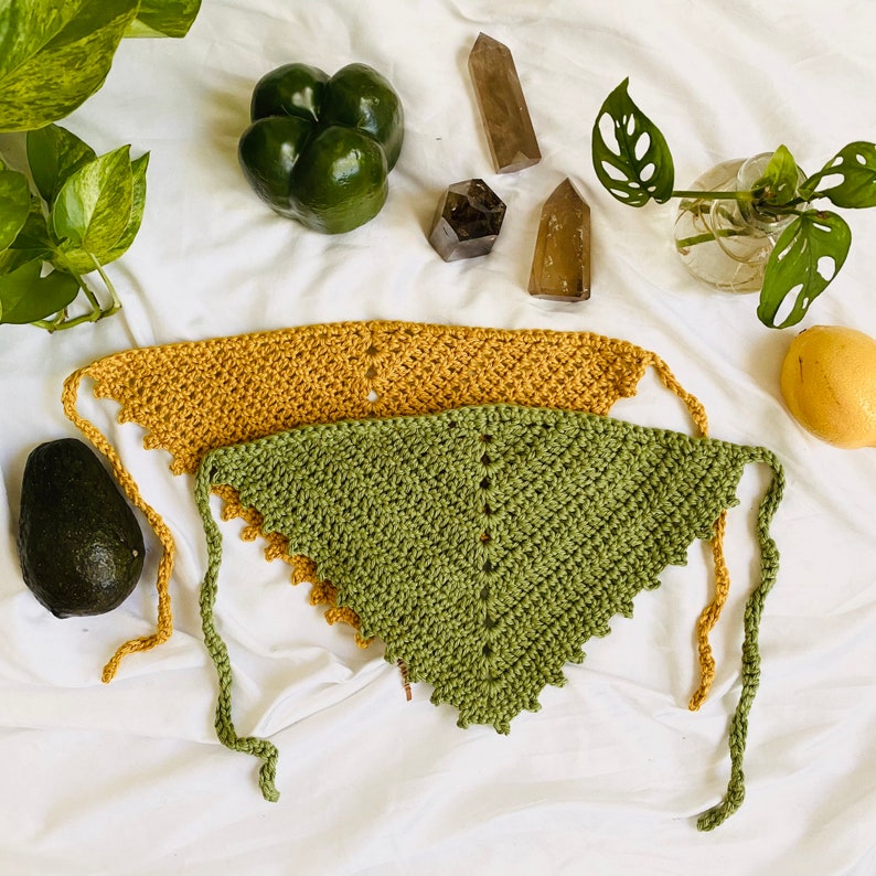 Puede incluir: Dos bufandas triangulares de crochet, una verde y otra amarilla mostaza, con bordes de flecos. Las bufandas est&aacute;n dispuestas sobre una superficie blanca con plantas verdes, aguacates y cristales.