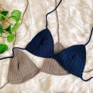 Pu&ograve; includere: Due top di bikini triangolari all'uncinetto, uno di colore marrone chiaro e l'altro di colore blu scuro. I top sono disposti su una superficie bianca e soffice.