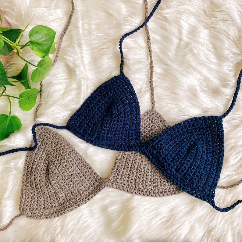 Pu&ograve; includere: Due top di bikini triangolari all'uncinetto, uno di colore marrone chiaro e l'altro di colore blu scuro. I top sono disposti su una superficie bianca e soffice.