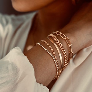 May include: A close-up of a person's wrist wearing five gold bracelets. The bracelets are stacked on top of each other and include a chain link bracelet, a thin gold band, a delicate chain with small charms, and a sparkly chain bracelet.