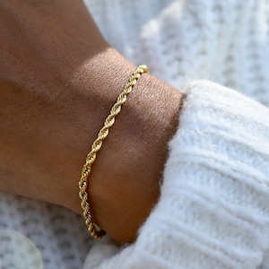 Op de afbeelding: Een goudkleurige armband met een touwketting. De armband wordt om een pols gedragen en de close-up toont het ingewikkelde gedraaide ontwerp. De armband is een eenvoudig maar elegant sieraad.