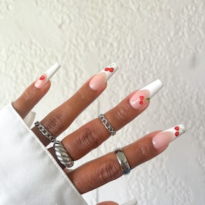 Könnte beinhalten: Nahaufnahme einer Hand mit langen, weißen, mandelförmigen Nägeln mit weißer Spitze und einem Kirsch-Design. Die Nägel sind mit einem hellrosa Lack lackiert. Die Hand trägt mehrere silberne Ringe.