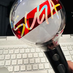 Könnte beinhalten: Ein klarer, kugelförmiger Leuchtstab mit dem Wort "SUGA" in holografischer Schrift über einem roten Band. Der Griff ist schwarz mit zwei silbernen Knöpfen. Der Hintergrund umfasst eine weiße Laptop-Tastatur.