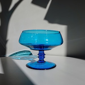 May include: A vibrant blue glass dessert bowl with a wide, shallow top and a detailed stem. The bowl sits on a white surface, with shadows cast across it. The glass has a glossy finish.