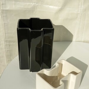 May include: A black, glossy ceramic vase with a unique, angular design stands next to a smaller, off-white ceramic container. Both pieces have a modern, geometric aesthetic, suitable for home decor.