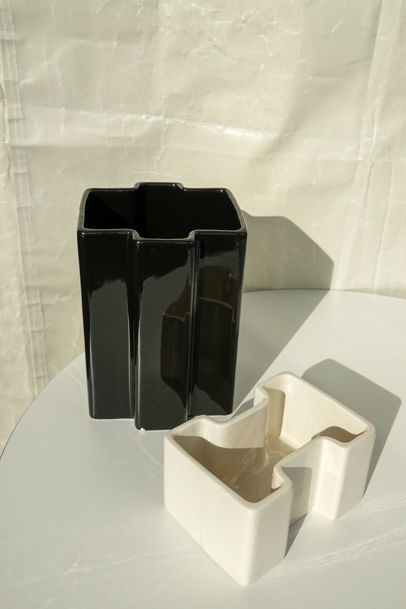 May include: A black, glossy ceramic vase with a unique, angular design stands next to a smaller, off-white ceramic container. Both pieces have a modern, geometric aesthetic, suitable for home decor.