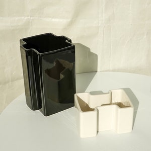 May include: Two geometric ceramic vases. One is a tall, glossy black, rectangular shape with a unique top. The other is a smaller, off-white, angular design. Both vases are on a white surface.