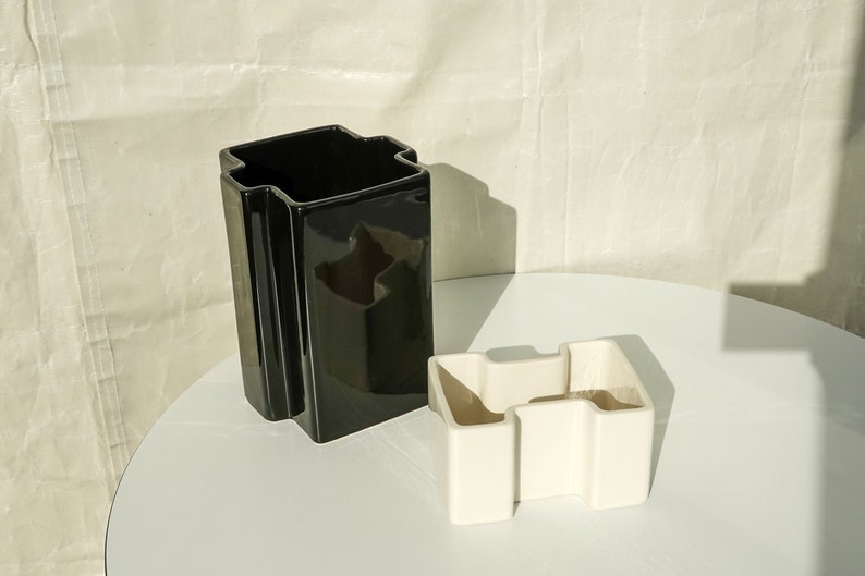May include: Two geometric ceramic vases. One is a tall, glossy black, rectangular shape with a unique top. The other is a smaller, off-white, angular design. Both vases are on a white surface.