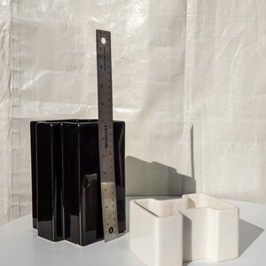 May include: A black ceramic vase with a fluted design, approximately 10 inches tall, next to a white ceramic container and a metal ruler. The vase has a glossy finish. The white container has a geometric shape.