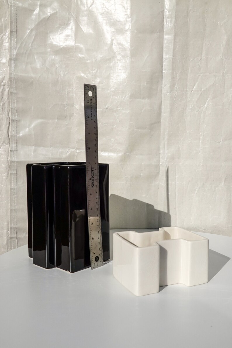 May include: A black ceramic vase with a fluted design, approximately 10 inches tall, next to a white ceramic container and a metal ruler. The vase has a glossy finish. The white container has a geometric shape.