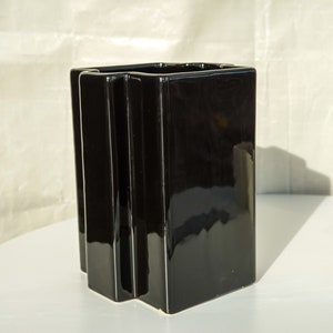 May include: A glossy black rectangular vase with a geometric design. The vase has a square opening and vertical, ridged details on the front. It is displayed on a white surface.