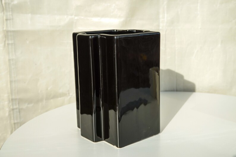 May include: A glossy black rectangular vase with a geometric design. The vase has a square opening and vertical, ridged details on the front. It is displayed on a white surface.
