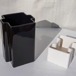 May include: Two ceramic vases, one black and one white, are displayed on a white surface. The black vase is tall and rectangular with a unique, stepped top. The white vase is smaller, with a similar design.