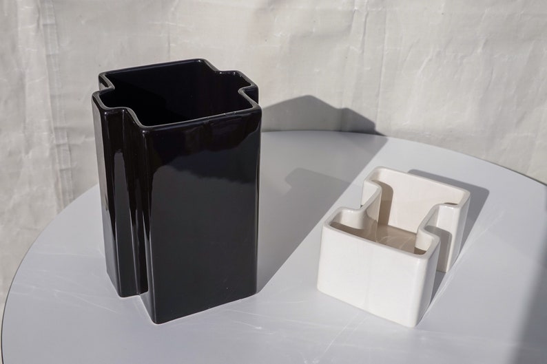 May include: Two ceramic vases, one black and one white, are displayed on a white surface. The black vase is tall and rectangular with a unique, stepped top. The white vase is smaller, with a similar design.