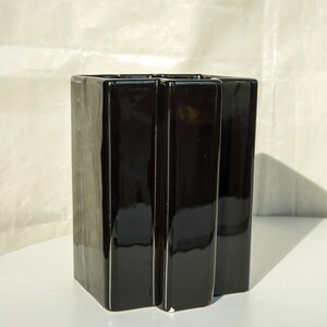 May include: A glossy black ceramic vase with a geometric design. The vase has a rectangular shape with vertical, slightly curved panels. The vase is on a white surface.