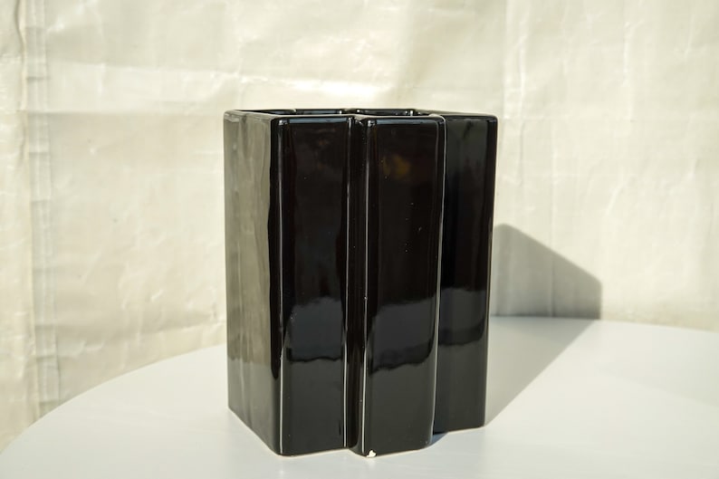 May include: A glossy black ceramic vase with a geometric design. The vase has a rectangular shape with vertical, slightly curved panels. The vase is on a white surface.