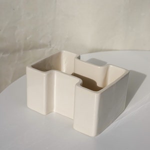 May include: Off-white ceramic container with a unique, geometric design. The container has a zig-zag shape with three open compartments. The surface is smooth and glossy, and the piece sits on a white surface.