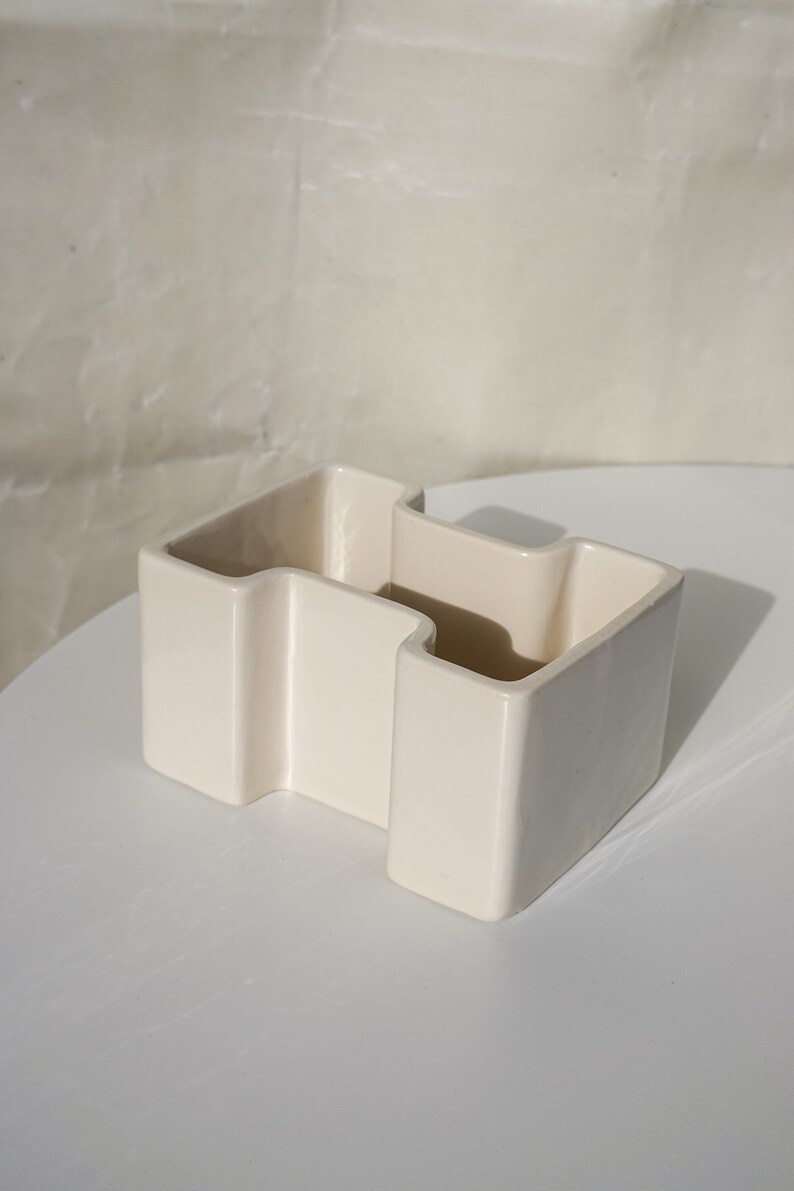 May include: Off-white ceramic container with a unique, geometric design. The container has a zig-zag shape with three open compartments. The surface is smooth and glossy, and the piece sits on a white surface.