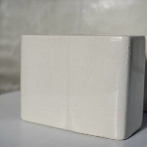 May include: Two off-white, rectangular ceramic vases. The vases have a slightly textured surface and a minimalist design. The soft lighting highlights the subtle imperfections and the clean lines of the vases, suggesting a modern aesthetic.
