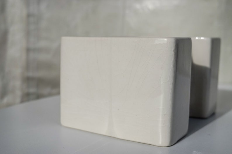 May include: Two off-white, rectangular ceramic vases. The vases have a slightly textured surface and a minimalist design. The soft lighting highlights the subtle imperfections and the clean lines of the vases, suggesting a modern aesthetic.