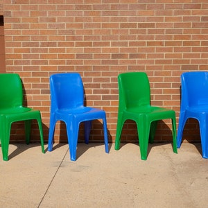 May include: Four plastic chairs in a row, alternating green and blue. The chairs are set against a red brick wall and a brown door. The chairs are made of a shiny plastic material.