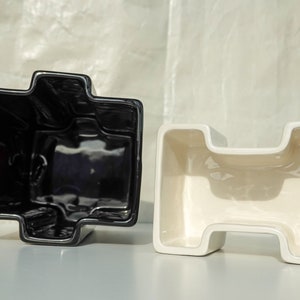 May include: Two ceramic planters, one black and one cream, are displayed. Each planter has a unique, geometric shape with a cross-like design. The planters are on a white surface, with a soft, textured background.