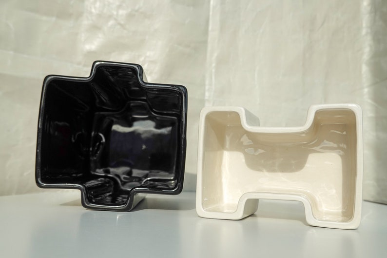 May include: Two ceramic planters, one black and one cream, are displayed. Each planter has a unique, geometric shape with a cross-like design. The planters are on a white surface, with a soft, textured background.