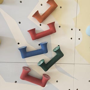 Puede incluir: Una selección de presas de escalada coloridas en naranja, rojo, azul y verde, montadas en una pared de escalada de madera clara. Las presas tienen forma de la letra "J" y están diseñadas para un agarre fácil.