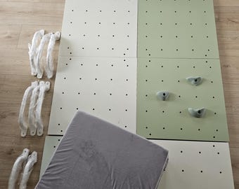 Puede incluir: Un conjunto de pared de escalada desmontado con paneles verdes y blancos, detalles de puntos negros y presas de escalada grises. Incluye una colchoneta gris acolchada y varios agarres plateados, todo sobre un suelo de madera clara.