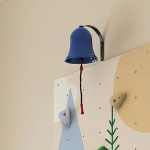 Pode incluir: Um sino azul com um suporte preto está montado na parede. Uma corda vermelha e preta pende do sino. A parede tem um design de parede de escalada com pegas coloridas.
