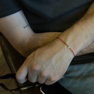 May include: A red string bracelet with a small gold bead charm. The bracelet is worn on a person's wrist.