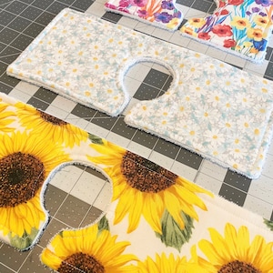May include: A selection of fabric sink skirt covers with floral designs. One features sunflowers, another has white daisies on a pale blue backdrop, and others display colourful flower arrangements. Each cover has a curved cutout.
