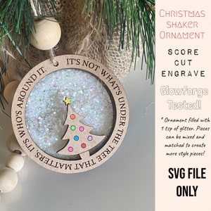 May include: Wooden Christmas shaker ornament with a tree design and glitter inside. The ornament has the text "IT'S NOT WHAT'S UNDER THE TREE THAT MATTERS, IT'S WHO'S AROUND IT." The tree has colorful dot ornaments.