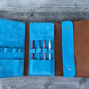 May include: Open, turquoise blue leather crochet hook case with brown leather accents. The case is open, revealing several crochet hooks in various sizes and colors. The case is designed to hold crochet hooks and other small tools.