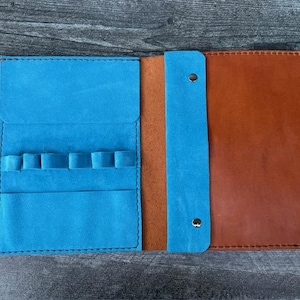 May include: Open leather pen case in shades of blue and brown. The case has a blue interior with multiple pen slots and a brown exterior. The case is open, revealing the interior and exterior colors. The case is on a wooden surface.