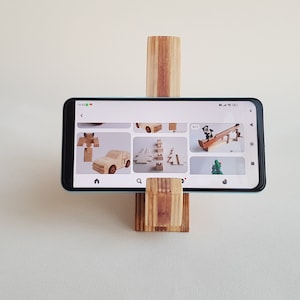 Op de afbeelding: Een houten telefoonstandaard met een lichtbruine afwerking houdt een smartphone vast die een scherm met afbeeldingen van houten speelgoed weergeeft. De telefoonstandaard is gemaakt van meerdere stukken hout die op elkaar zijn gestapeld en aan elkaar gelijmd zijn. De telefoonstandaard is een eenvoudig en functioneel ontwerp.