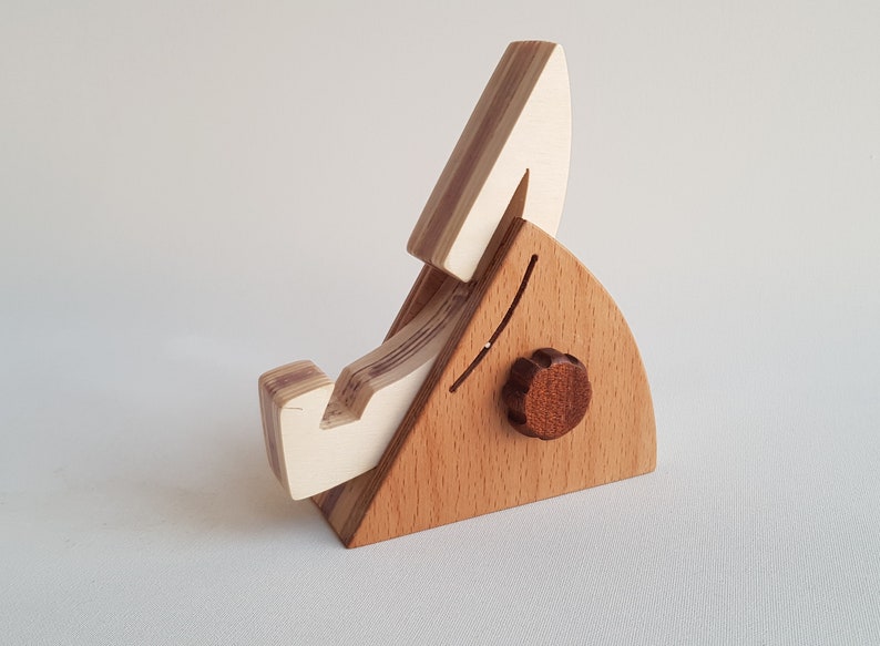 Op de afbeelding: Een houten telefoonstandaard met een wit en bruin gestreept ontwerp. De standaard heeft een kleine knop aan de zijkant en een gebogen gleuf om een telefoon vast te houden.