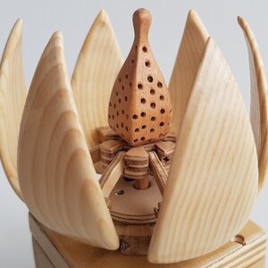 May include: A wooden sculpture of a flower with a detailed, intricate design. The flower is made of light wood and has a brown centre with small holes. The flower is sitting on a wooden base.