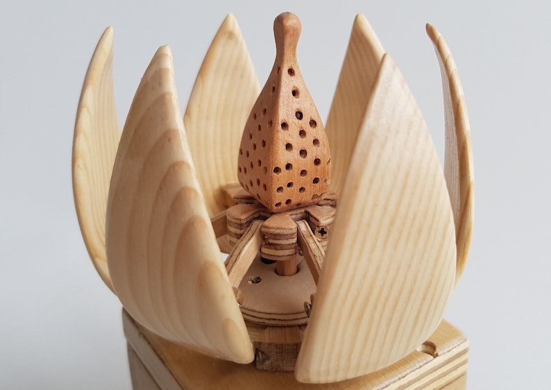 May include: A wooden sculpture of a flower with a detailed, intricate design. The flower is made of light wood and has a brown centre with small holes. The flower is sitting on a wooden base.