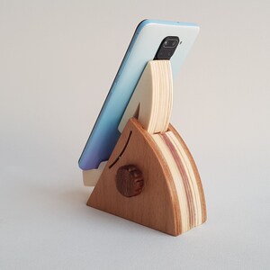 Op de afbeelding: Een houten telefoonstandaard in de vorm van een driehoek met een lichtblauwe telefoon erop. De standaard is gemaakt van licht en donker hout.
